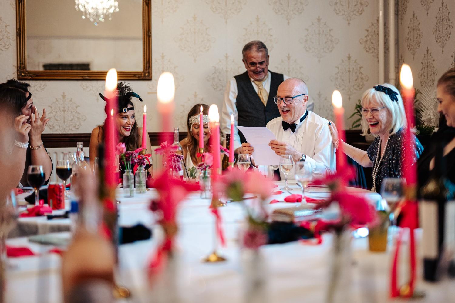 Mixed age group party of colleagues seated around a table with pink candles. They are all dressed up in 1920's themed clothing, and are participating in a murder mystery evening. Murder mystery events are a popular choice as an added activity at your Party House.