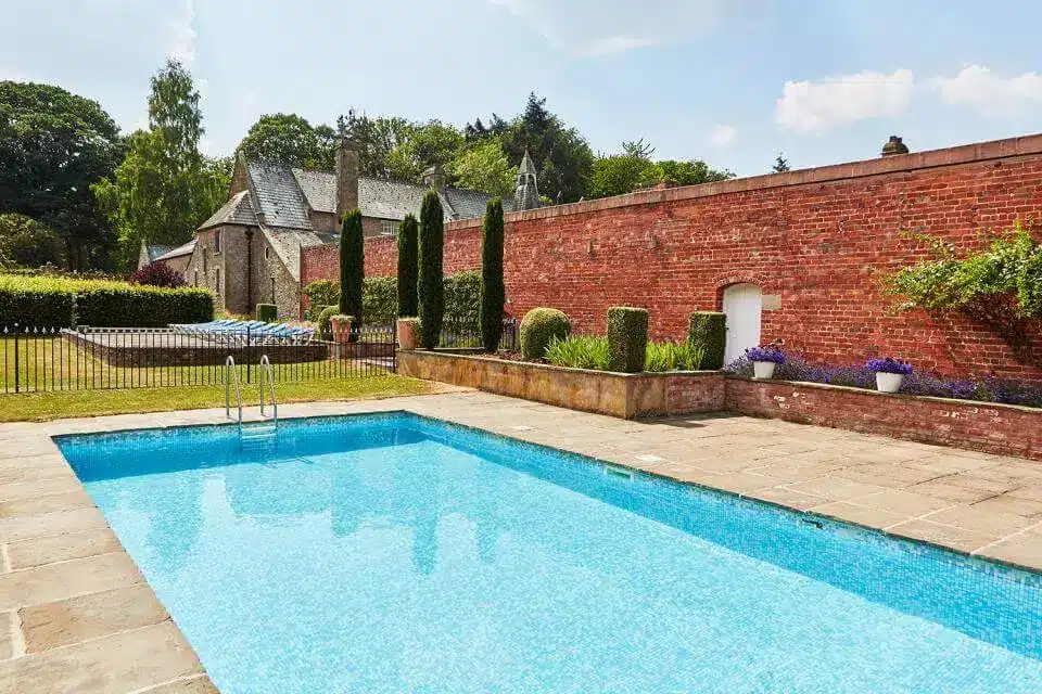 Nright blue swimming pool next to a red brick wall with some grass and trees in the distance